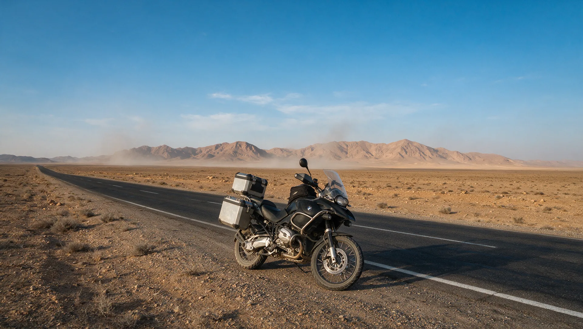 Beladenes Reiseenduro-Motorrad am Rand einer leeren Asphaltstraße durch die weite, karge Hochwüste des iranischen Plateaus, ferne braune Berge unter klarem blauem Himmel